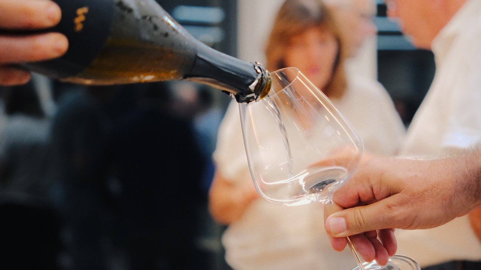Wein wird in ein Glas eingeschenkt, dahinter unscharf die Gäste.